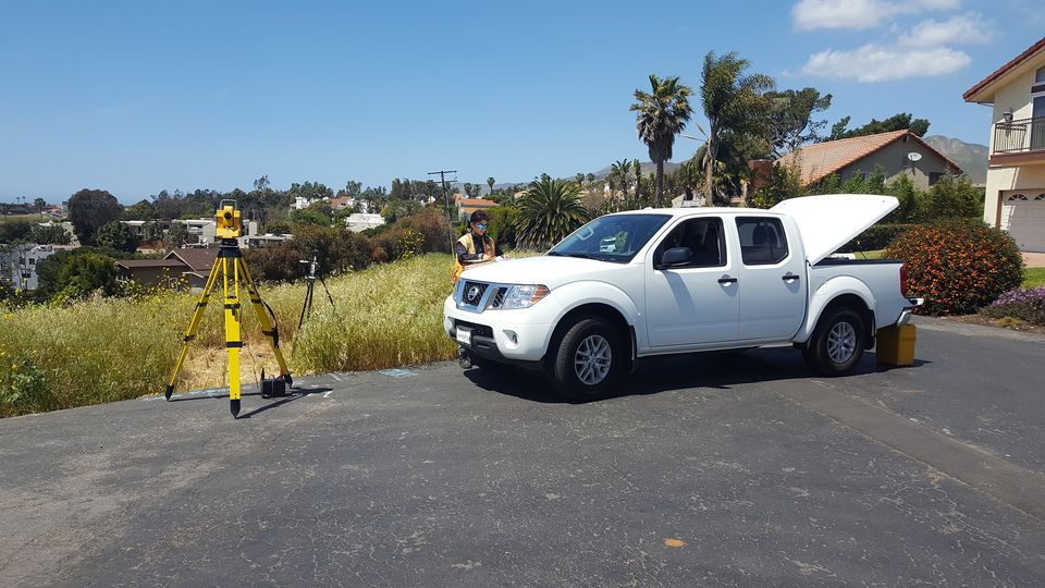 Image shows a land surveyor and story pole worker standing by a work truck preparing to start a job. a surveying instrument stands on a tri pod mount